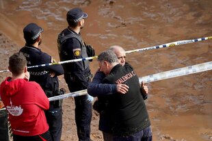 Menschen liegen sich weinend in den Armen, dahinter stehen Polizisten hinter Absperrband, das die Menschen von schlammigen Wassermassen trennt.