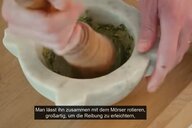 Zwei Hände bewegen einen Stößel in einem Mörser aus Marmor.