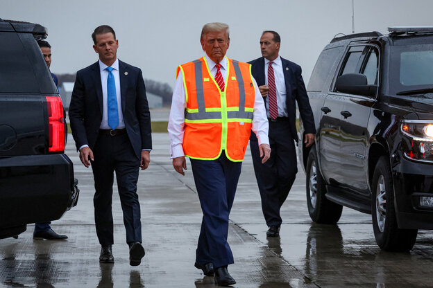 Donald Trump geht in orangener Warnweste mit zwei anzugtragenden Männern zwischen Autos entlang.