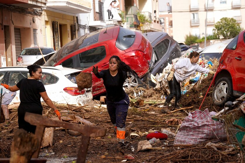 Drei Menschen mit langen Haaren stehen mit Gummistiefeln in den Trümmern auf einer Straße, auf der mehrere Autos vom Hochwasser durch die Gegend gespült wurden