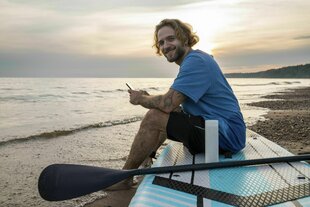 Sonnenuntergang am Meer: Junger sportlicher Mann sitzt auf einem Paddelboard mit Paddel am Strand und hat sein Smartphone in der Hand.