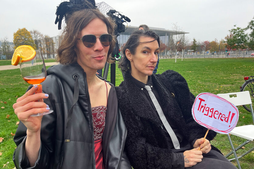 Zwei Transfrauen mit Aperolglas und Schild TRIGGERED in den Händen