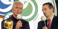 zwei Männer mit Mikros vor einer Wand mit dem Logo der Fußball-Weltmeisterschaft 2006, vor ihnen der WM-Pokal. Die Männer sind Franz Beckenbauer und Wolfgang Niersbach