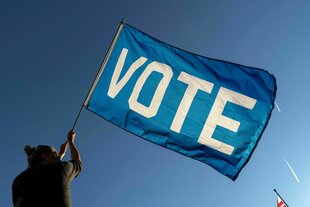 Ein Mensch von hinten ist zu sehen, er steht vor strahlend blauem Himmel und hält eine riesige blaue Flagge, auf der das Wort "Vote" steht