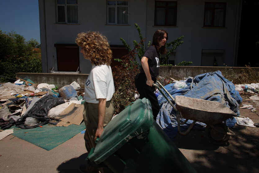 Zwei Frauen sind vor einem Haus zwischen Müllsäcken mit Schubkarre und Mülltonne zugange