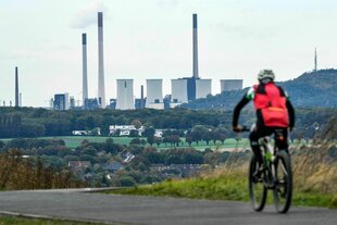 Ein Mann fährt Fahrrad in Richtung eines Kohlekraftwerks mit vielen Schloten und Kühltürmen in grüner Landschaft