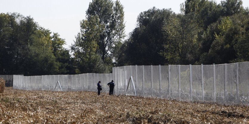 Zaun führt über ein Feld und überragt bei weitem zwei Menschen, die davor stehen