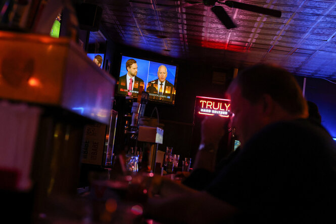 Personen sitzen am Tresen einer Bar, an der Wand hängt ein Bildschirm, auf dem die Gesichter von Vance und Walz zu sehen sind