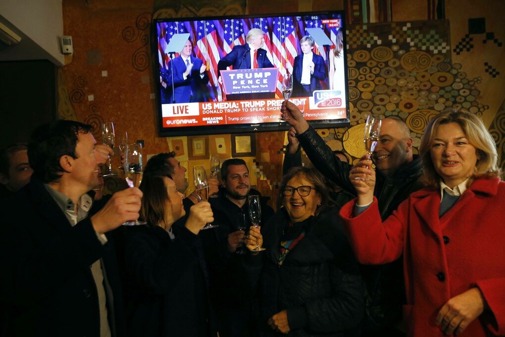 Menschen feiern unter einem Fernseher, der Trump zeigt.