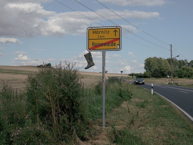 Ein Gummistiefel hängt am Ortseingangsschild von Hainewalde, auf der Landstraße fährt ein Auto