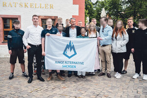 Eine Gruppe von 17 Personen, überragend männlich gelesen, stehen vor einem miitelalterlichen Haus auf welchem in Gold geschrieben steht 