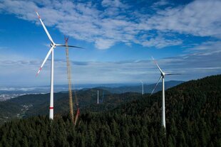 Ein Kran zieht das dritte Rotorblatt einer neuen Windkraftanlage auf der Holzschlägermatte am Schauinsland hoch während daneben eine kleinere Windkraftanlage und im Hintergrund eine weitere im Bau befindliche Windkraftanlage zu sehen sind.