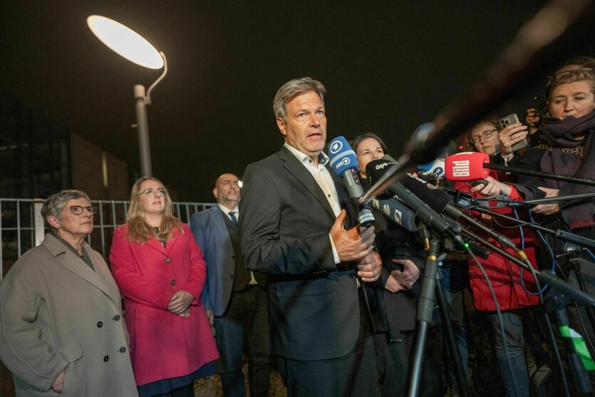 Robert Habeck und Annalena Baerbock stehen vor dem Kanzleramt, umringt von Journalisten, die ihnen die Mikros entgegen strecken. Die Szenerie wird von einer großen Lampe beleuchtet.