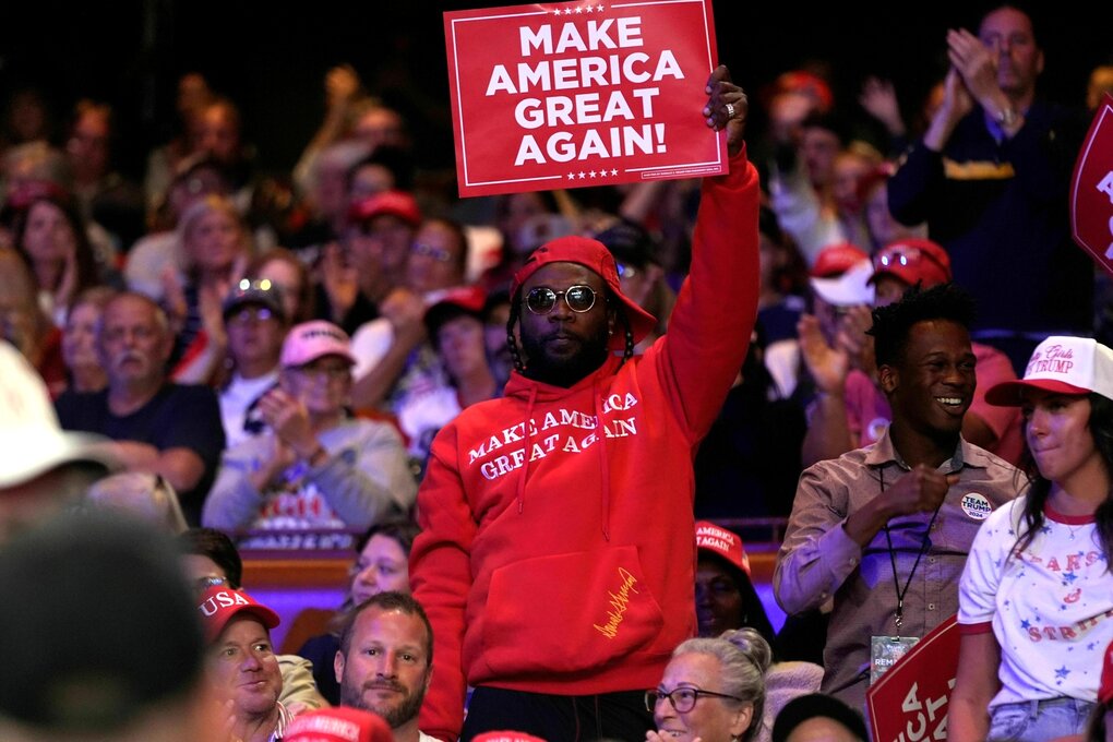 Ein Afroamerikaner mit rotem Basecap und Hoodie mit der Aufschrift: Make America great again, hält während einer Wahlkampfveranstaltung ein Schild hoch: Make America great again