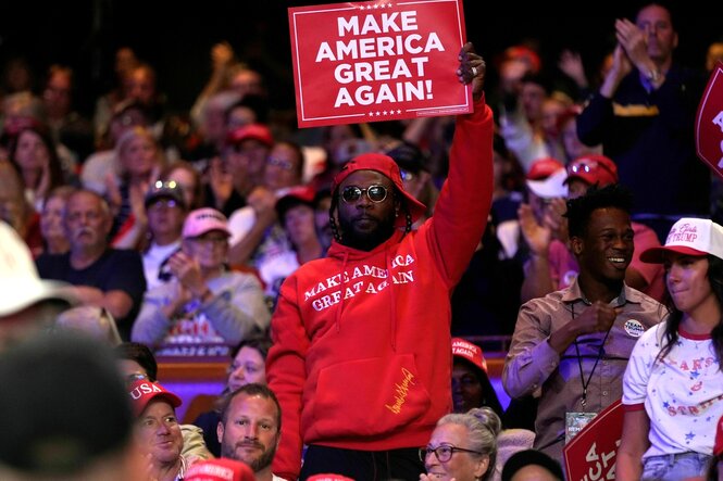 Ein Afroamerikaner mit rotem Basecap und Hoodie mit der Aufschrift: Make America great again, hält während einer Wahlkampfveranstaltung ein Schild hoch: Make America great again