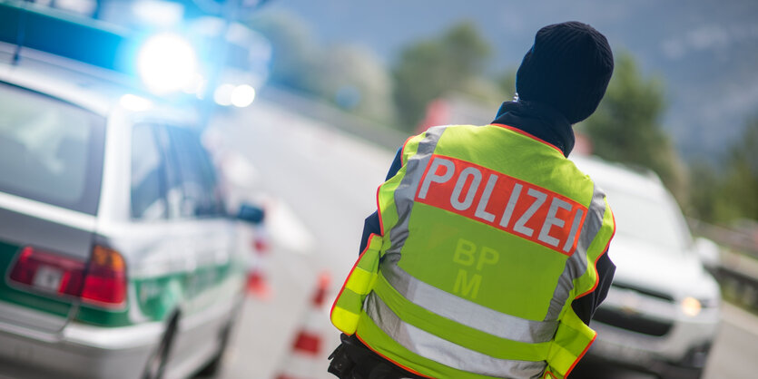 Ein Polizist steht mit dem Rücken zum Betrachter. Er hat eine Warnweste an, damit er von den Autos gut gesehen wird.