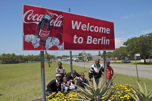 Eine Gruppe Kinder sitzt in Südafrika unter einem Ortschild mit Coca Cola-Werbung.