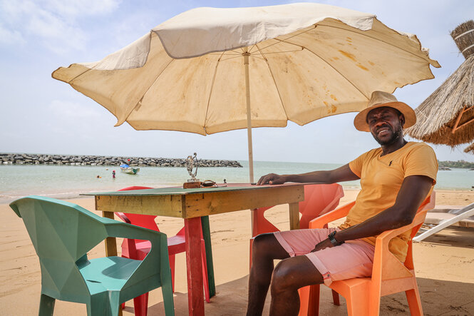 Ein Mann sitzt lässig unter einem Sonnenschirm am Strand an einem Tisch