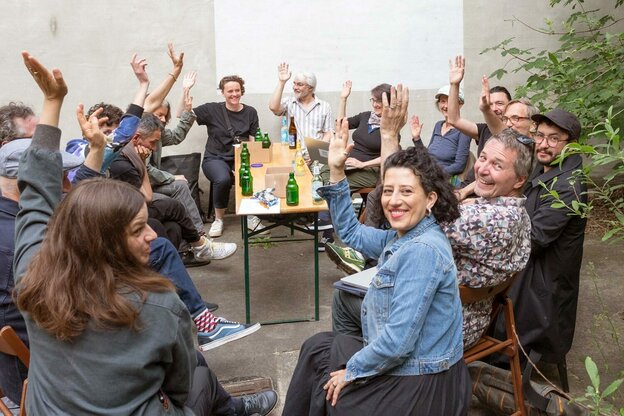 Eine Gruppe von Klangkünstler:innen sitzen in einem Projektraum im Kreis, sie winken in die Kamera