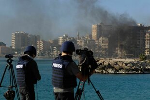 Zwei Kameramänner mit Pressewesten stehen am Wasser. Am anderen Ufer sind Gebäude zu sehen, aus denen schwarzer Rauch aufsteigt.