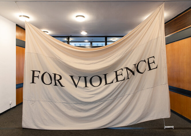 Ein weißes Tuch hängt in einem Ausstellungsraum von der Decke. Darauf sind in Versalschrift "FOR VIOLENCE" gedruckt.