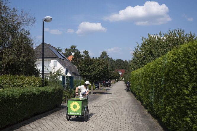 Eine Frau mmit einem Lastenfahrrad mit Grünenaufkleber dreht sich auf einem Weg zwischen kleinen Häusern um