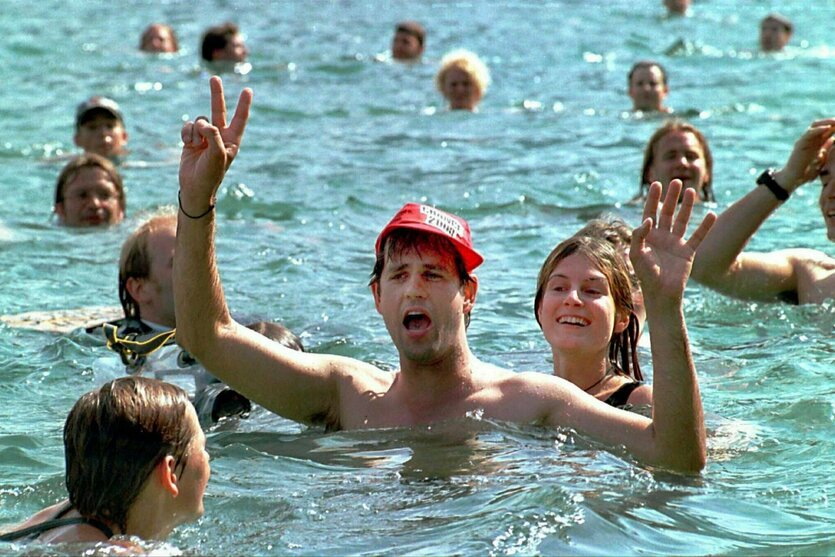 Christoph Schlingensief beim Protest-Baden in einem See, umgeben von Mitbadenden. Er trägt eine rote Kappe mit Aufschrift: Chance 2000