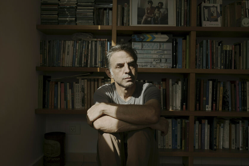 Etgar Keret sitzt von Sonnelicht beschienen mit verschränkten Armen auf dem Boden vor einem Bücherregal und blickt nachdenklich. Das Foto entstand wenige Wochen nach dem 7. Oktober 2023