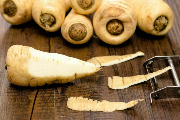 Pastinaken liegen auf einem Holztisch, eine Pastinake wurde schon angeschält, der Schäler ist im Anschnitt zu sehen