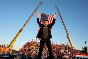 Wahlkampfveranstaltung von Donald Trump, Menschenmasse auf einer Tribüne im Hintergrund. Elon Musk steht breitbeinig vor einer rießigen US-Flagge, die an zwei Baukränen befestigt ist - auf der Bühne und reckt die Arme nach obn