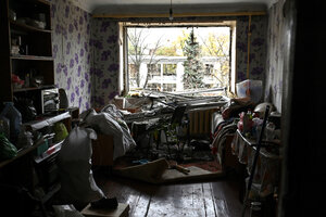 Ein Zimmer dessen Fensterscheibe zerstört ist, Scherben Fensterrahmen sind auf einen Schreibtisch gefallen, es herrscht Chaos. Der Blick nach draussen zeigt einen Tannnbaum und ein flaches Gebäude, dessen Fenster auch zerstört und beschädigt sind