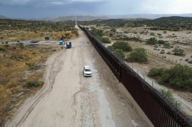 Eine Zaun ewig langer brauenr Zaun mit Stacheldraht bestückt teilt eine Prärielandschaft. Auf der staubigen Piste vor dem Zaun fährt ein heller Truck, weiter hinten steht ein Container, flankeirt von zwei Dixie-Toiletten