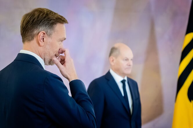 Christian Lindner steht im Vordergrund nahe bei einer deutschen Fahne, die ins Bild reinragt. Er fasst sich an die Nase. Im Hintergrund sieht man Olaf Scholz