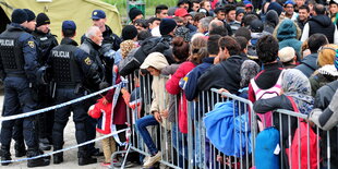 Dutzende Menschen warten an einem Zaun, dahinter Polizisten und ein Zelt.