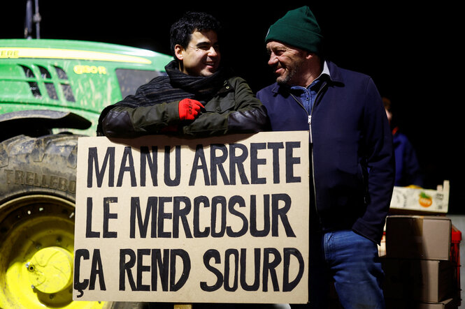 Zwei Bauern stehen hinter einem Schild. Darauf steht: Manu, stopp Mercosur, das macht einen taub.“ Gemeint ist der französische Präsident Emmanuel Macron