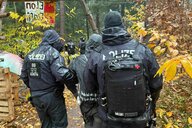 Polizisten führen eine Person ab, sie laufen auf einem Waldweg, es regnet