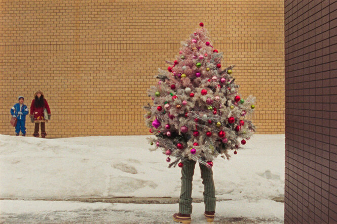 Zwei Kinder stehen im Winter vor einer gelben Hauswand. Sie schauen auf einen Weihnachtsbaum, aus dem die Beine eines Menschen herausschauen.