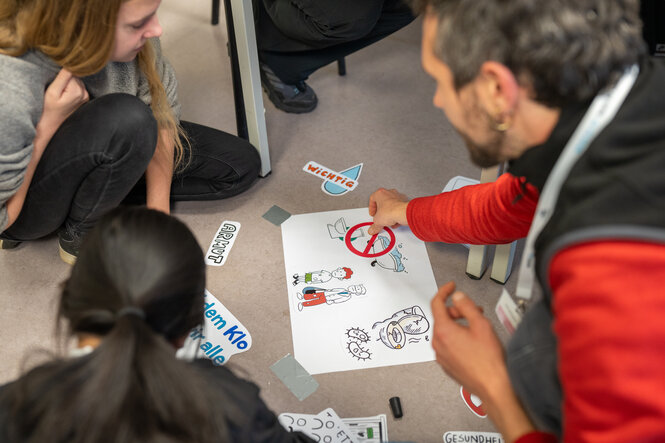 Kinder sitzen bei einem Workshop um ein Blatt Papier, auf dem verschiedene Toiletten gezeichnet sind