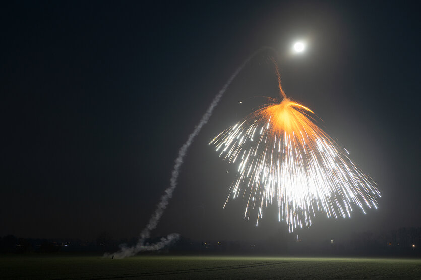 Ein Feuerwerk am Nachhimmel. Orangende und weiße Funken fügen sich zu einem Leuchtregen zusammen. Im Hintergrund ist die Rauchspur des Feuerwerks zu sehen.