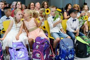 Festlich gekleidete Schulkinder sitzen in Stuhlreihen, vor ihnen stehen ihre Ranzen auf dem Boden, einige Kinder halten eine Sonnenblume in der Hand