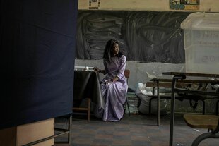 Eine Frau in einem violetten Kleid sitzt in einem dunklen Raum zwischen einfachen Tischen, Stühlen und Kunststoffbehältern