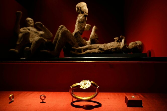 Vitrine im Vordergrund mit zwei Ringen und einem Armreif auf rotem Untergrund, im Hintergrund die Gipsabdrücke von Opfern des Vesuvausbruchs