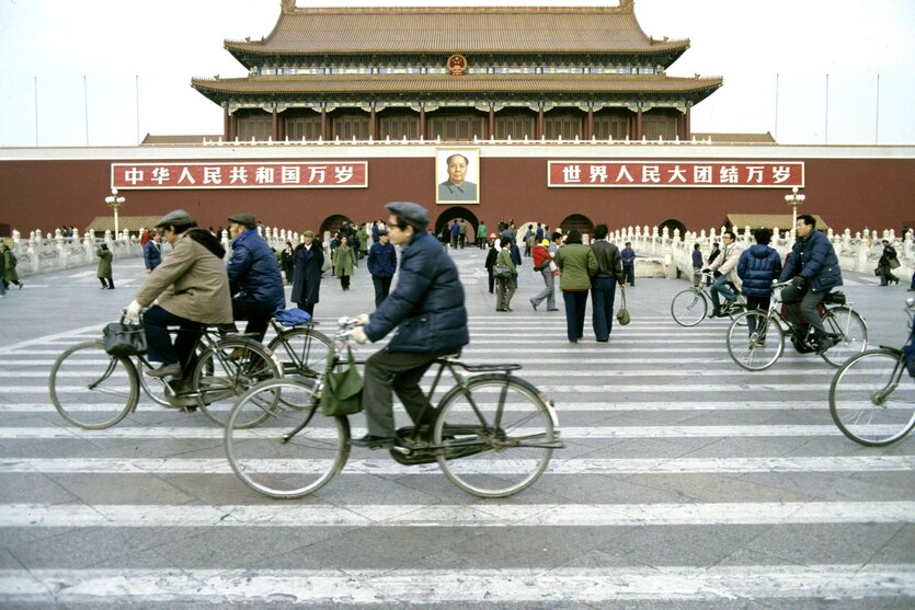 Menschen radeln in den 80er Jahren am Tiananmen Platz mit dem Porträt von Mao, vorbei