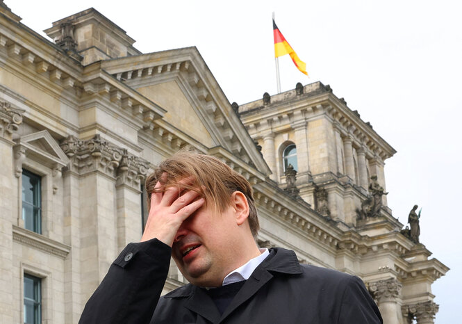 Ein Mann steht vor dem Reichtstag und hält sich eine Hand vor das Gesicht