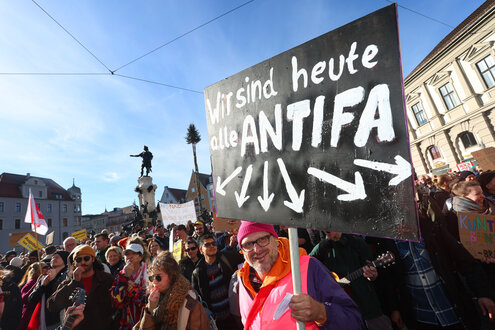 Bei einer Demo gegen rechts steht ein Demonstrant mit einem Plakat, auf dem steht: Wir sind heute alle Antifa. Pfeile zeigen auf die Demonstrationsteilnehmer.