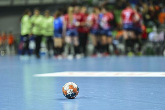 Ein Handball liegt im Vordergrund auf dem Boden. Im Hintergrund sieht man unscharf einen Pulk aus Spielerinnen