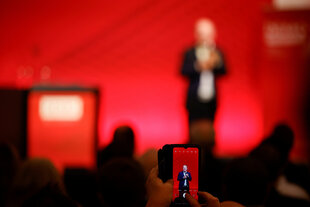 Olaf Scholz steht auf einer Bühne vor rotem Hintergrund, im Vordergrund ein Smartphone, auf dessem screen er scharf zu sehen ist