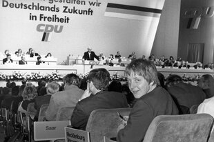 Angela Merkel blickt über die Schulter zum Fotografen, sie ditzt im Publikum auf dem CDU-Parteitag, am Rednerpult steht Helmut Kohl, Archivfoto von 1991