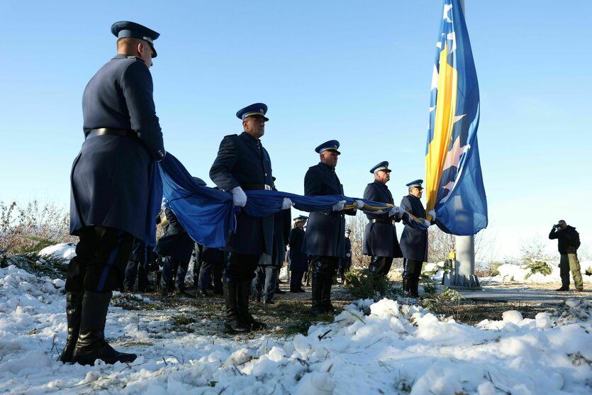 Männer in Uniform stehen vor einem Fahnenmast mit der bosnischen Flagge, deren unteren Teil sie gemeinsam in den Händen halten, damit sie den Boden nicht berührt