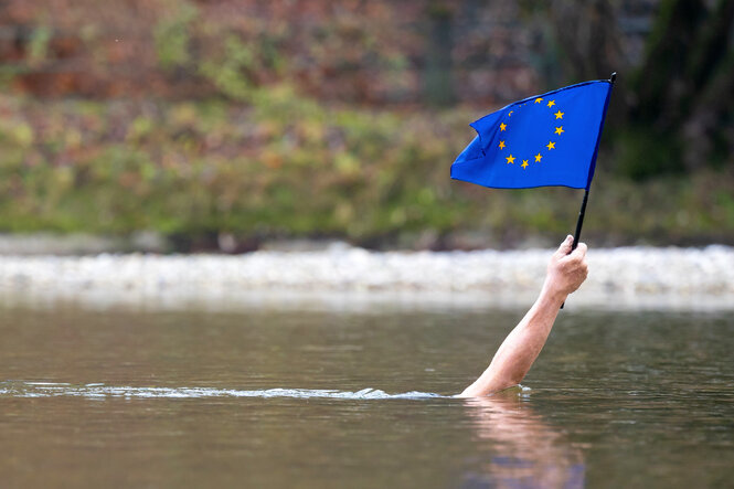 Ein menschlicher Arm guckt aus einem Fluss, in der Hand eine kleine Europa-Flagge